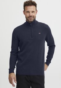 Marineblå hettegenser med glidelås i myk strikket stoff, med høy krage, ribbestrikkede mansjetter og en liten logo på brystet.