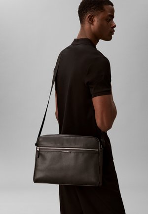 Man draagt zwarte polo en zwarte textuur leren schoudertas met voorvak met ritssluiting en Calvin Klein-logo op de band.