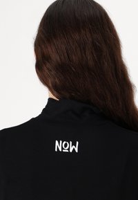 Svart skjorte med høy krage og en hvit "NOW" logo trykket på baksiden i en fet, leken skrifttype. Myk stofftekstur.