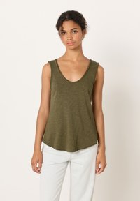 Giovane donna con capelli ricci che indossa un top verde oliva senza maniche e pantaloni chiari, in piedi davanti a uno sfondo chiaro uniforme.