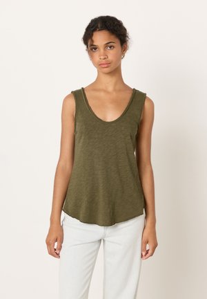 Nuori nainen kiharahiuksisena, yllään hihaton oliivinvihreä paita ja vaaleat housut, seisomassa yksinkertaisen vaalean taustan edessä.