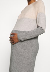 Gravid person som bär en lång, färgblockerad klänning i beige och grått, som varsamt håller om sin mage med båda händerna.