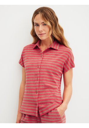 Woman with wavy blonde hair wearing a short-sleeve, button-up shirt with red and beige horizontal stripes, hand in matching striped pants pocket.