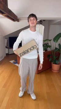 Rjav pravokoten škatla z napisom "zalando" natisnjenim s poudarjenimi črnimi črkami, ki jo drži oseba v beli srajci z dolgimi rokavi in sivo črtastih hlačah.