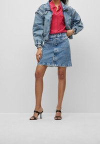 Jeansjacke mit gepufften Ärmeln, kombiniert mit einem rosa Hemd und einem hellblauen Jeansrock mit vorderen Knöpfen. Schwarze Absatzsandalen.