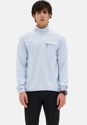 Giovane uomo che indossa un pullover azzurro chiaro con zip a quarto e tasca sul petto, e pantaloni sportivi neri, in piedi su uno sfondo semplice.