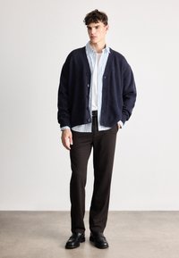 Tengerészkék kardigán gombos elülső résszel, puha textúrával és bordázott szélekkel, világoskék csíkos ing felett, sötét nadrág és fekete cipő kombinációjában.