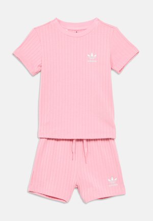 Lyserød ribbet Adidas t-shirt med korte ærmer og matchende shorts med hvide Adidas-logoer på brystet og venstre ben, vist på en hvid baggrund.