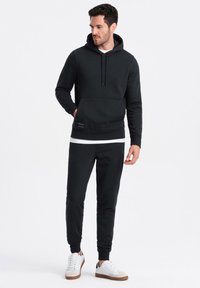 Sudadera negra con cordón y bolsillo canguro, combinada con pantalones jogger a juego. Textura suave, puños acanalados, acentuada con zapatillas blancas.