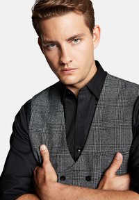 Szary kamizelka w kratę na czarnej koszuli, z podwójnym zapięciem, spiczastymi klapami i kieszeniami. Materiał wydaje się być teksturowany.