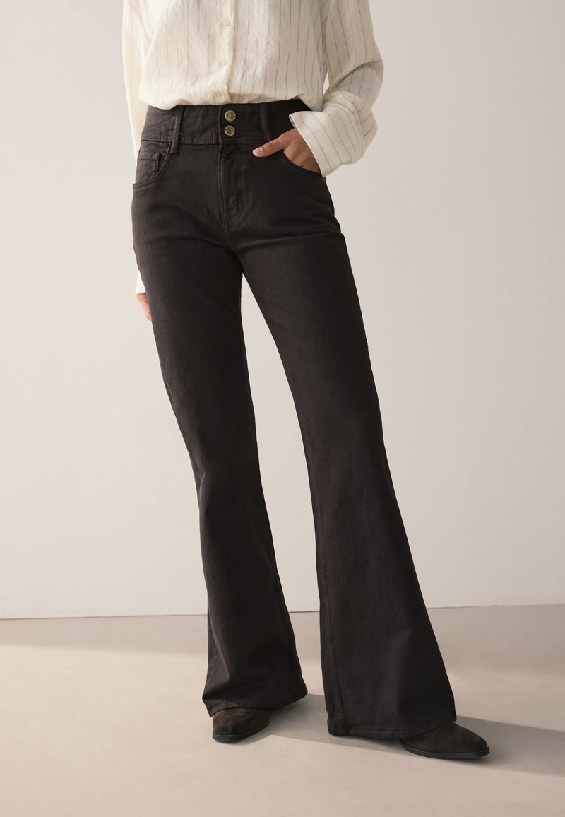 Personne portant un jean noir évasé taille haute avec fermeture double boutons et une chemise blanche à manches longues rentrée, debout avec une main dans la poche.