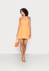 Vestido mini laranja sem mangas, com tecido texturizado, corpete franzido e saia rodada. Combinado com sapatos de salto laranja com tiras e uma bolsa pequena.