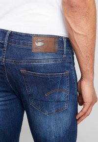 Mörkblå denimjeans med orange sömmar, en brun läderlapp på bakre midjebandet och en klassisk design med fem fickor.