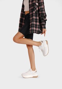 Scarpe da ginnastica bianche con pannelli testurizzati, suole spesse e accenti beige, abbinate a una camicia a scacchi nera e rosa e a pantaloni shorts neri.