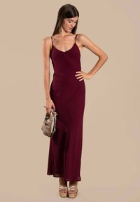 Bordeauxfarbenes, ärmelloses Maxikleid mit V-Ausschnitt, aus glattem Stoff gefertigt. Kombiniert mit goldenen Plateauschuhen und einer metallischen Clutch.