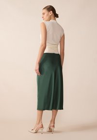 Jupe en satin vert avec un corsage crème ajusté, présentant un dos froncé, et associée à des escarpins beiges à bride arrière et une texture lisse.