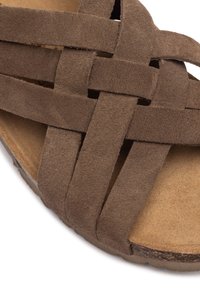 Sandalo in suede marrone con design a fascia intrecciata, dotato di suola piatta e sottopiede beige. La texture è morbida con una finitura naturale.