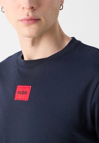 Sötétkék póló kerek nyakkal, elöl egy piros téglalap alakú folttal, amelyen feketével hímezve van a "HUGO" felirat.