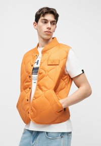Jeune homme portant une veste matelassée orange sur un t-shirt blanc avec un motif graphique et un jean bleu clair, posant avec une main dans la poche devant un fond blanc.