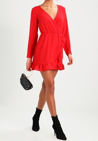 Vestido rojo envolvente con mangas largas, dobladillo con volantes y escote en V profundo. Combinado con un bolso de mano negro con un asa circular y acentos dorados.