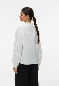 Witte blouse met gestructureerde stippen, ruches op de kraag en lange mouwen. Heeft een kanten rand langs de manchetten. Gepaard met zwarte broek.