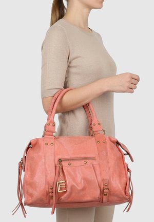 Femme en haut beige tenant un grand sac à main rose avec doubles poignées et une poche avant zippée avec un pendentif doré.