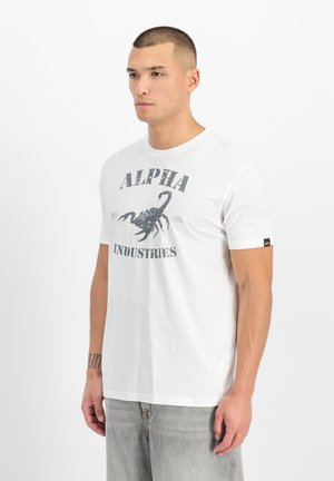 Homme aux cheveux courts portant un T-shirt blanc Alpha Industries avec un motif de scorpion noir et un jean gris, debout devant un fond blanc.