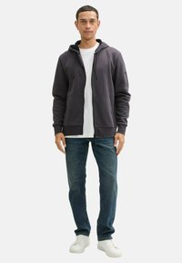 Felpa con zip di colore grigio scuro, realizzata in un materiale morbido, con due tasche frontali, cappuccio con cordino e polsini a costine, abbinata a jeans blu e sneakers bianche.