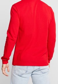 Maglietta rossa a maniche lunghe in cotone; presenta un design semplice con collo rotondo e maniche aderenti, abbinata a jeans in denim chiaro.