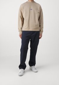 Sudadera beige de manga larga y cuello redondo, con un pequeño logo azul en el pecho. Combinada con pantalones azul marino y zapatillas blancas.