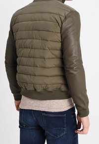 Man i olivgrön vadderad jacka med läderslag, beige tröja och mörkblå jeans, sedd bakifrån och står mot vit bakgrund.