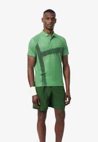 Grön pikétröja med svart abstrakt tryck, kort ärm och krage, kombinerad med mörkgröna shorts. Bomullsmaterialblandning.
