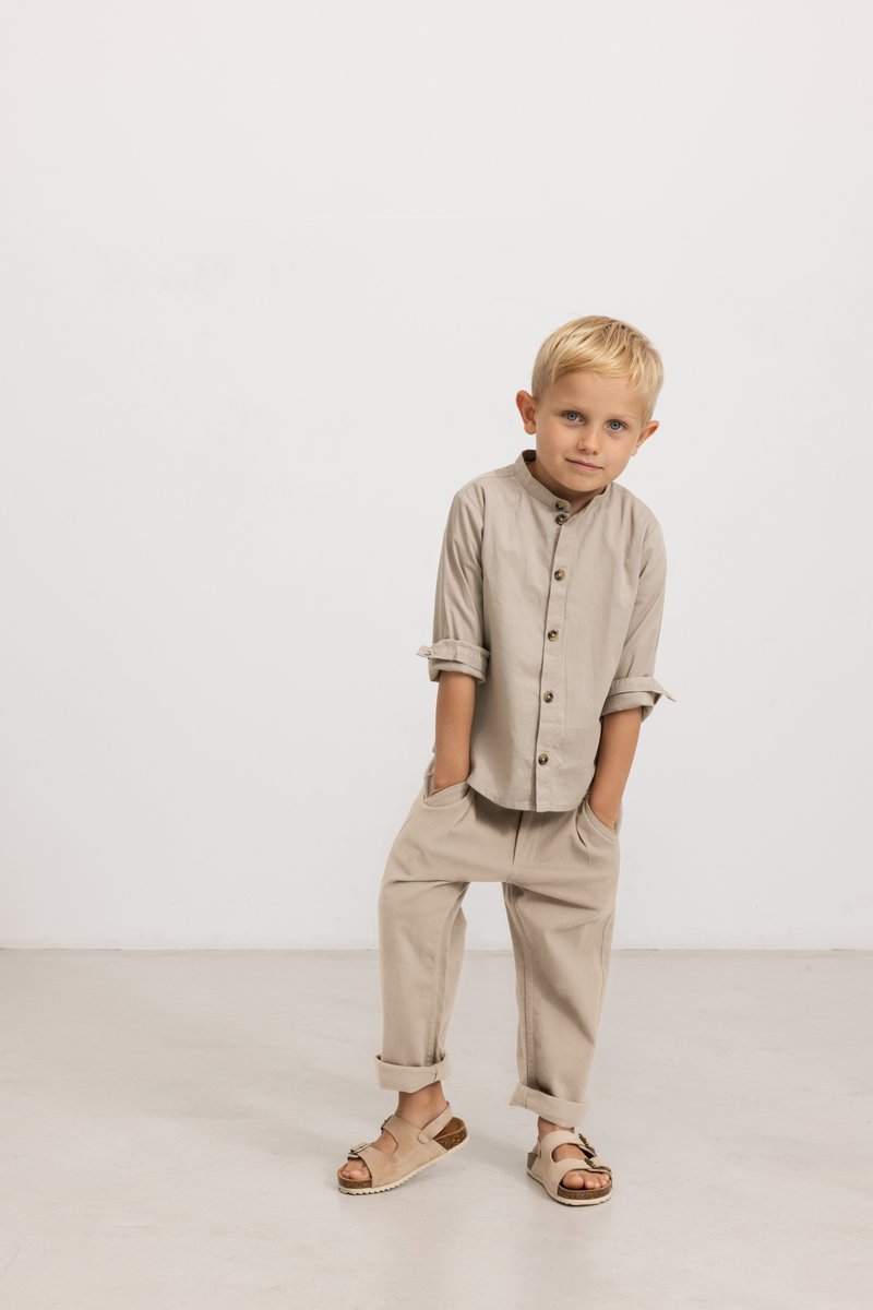Camicia di cotone beige con maniche arrotolate e bottoni di legno, abbinata a pantaloni beige larghi e sandali beige con un accento marrone.