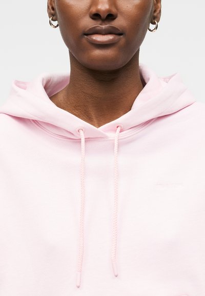 Inimese lähiplaan, kes kannab helesinist kapuutsiga dressipluusi, rõngaskujulisi kõrvarõngaid ja neutraalse tooniga huuli ühetoonilisel taustal.