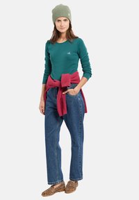 Langarmige teal shirt met een klein logo, bordeauxkleurige trui om de taille geknoopt, losse blauwe jeans en tan kleurige schoenen met luipaardprint. Groene gebreide beanie.