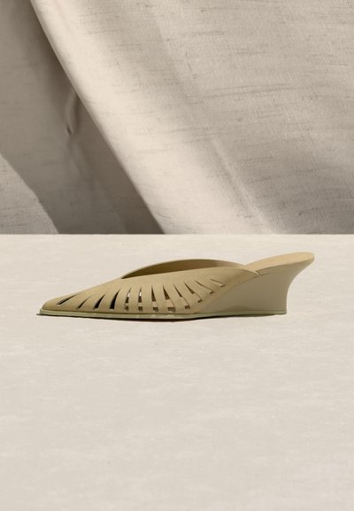 Μπεζ mule slip-on με τακούνι πλατφόρμα και σχέδιο κοψίματος στο πάνω μέρος, τοποθετημένο σε ουδέτερο υφασμάτινο φόντο και πάτωμα.