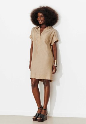 Femme avec une coiffure afro naturelle portant une robe beige à manches courtes et des sandales à plateforme noires, debout contre un mur blanc.