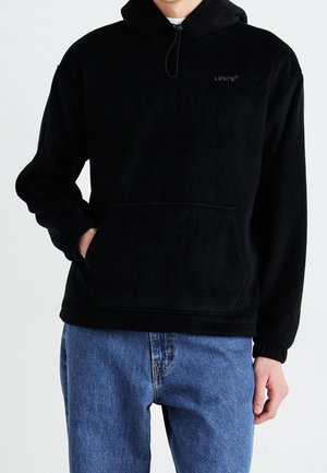 Henkilö pukeutuneena mustaan Levi's-fleecejakkuun, jossa on etutasku, ja sinisiin farkkuihin, käsi taskussa, yksinkertaista vaaleaa taustaa vasten.