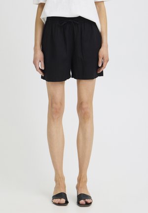 Person trägt schwarze Shorts mit elastischem Bund, weißes Top und schwarze offene Sandalen, steht vor einem schlichten hellen Hintergrund.