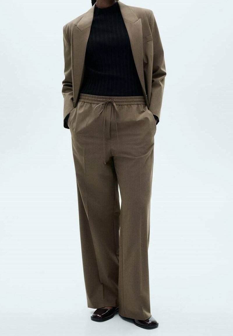 Personne portant un pantalon large marron sur mesure avec cordon, un blazer marron assorti, un col roulé noir côtelé et des chaussures sombres à enfiler.