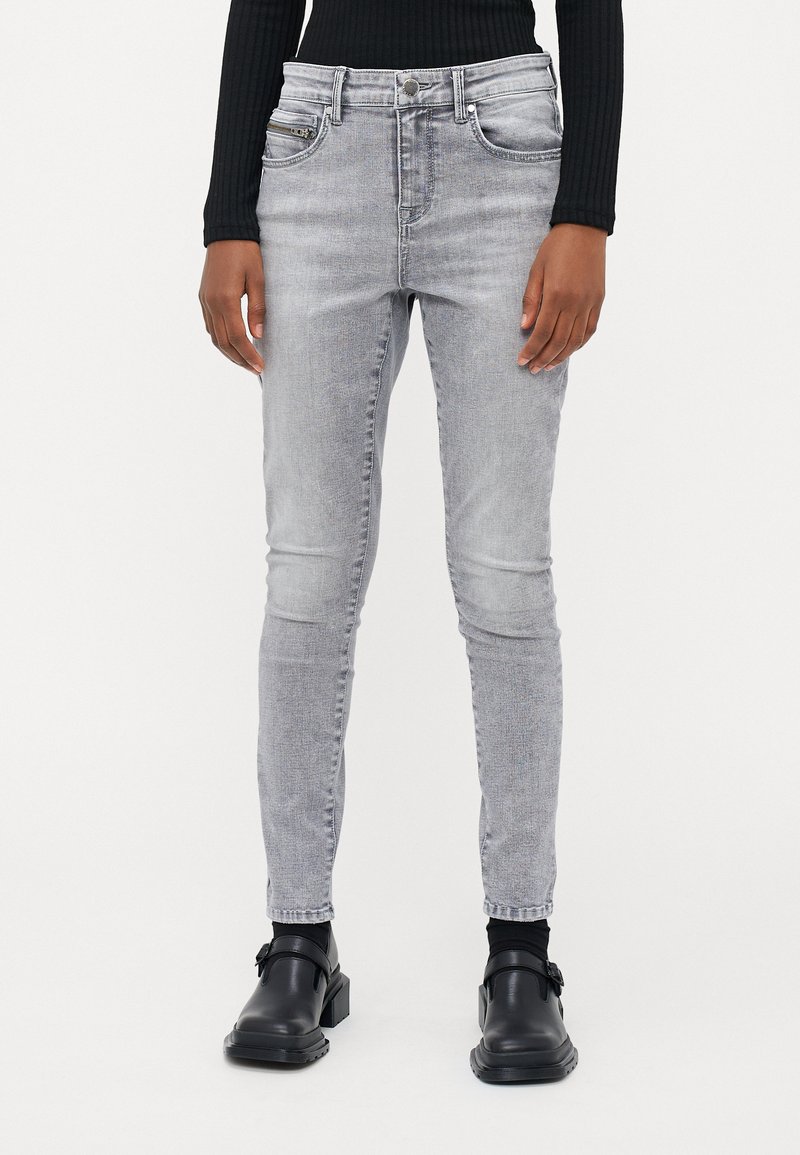 Világosszürke skinny farmernadrág sima textúrával, öt zsebes designnal és sárgaréz kiegészítőkkel. Fekete platform cipővel és testhezálló fekete toppal viselve.