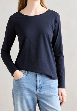 Person in dunkelblauem Langarmshirt und hellblauen Jeans, mit einer Hand in der Tasche, steht vor einem schlichten hellen Hintergrund.