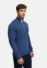 Blå stickad pullover med halvlång dragkedja i halsen, texturerade mönster, åtsittande ärmar och ribbade muddar, kombinerad med mörka byxor.