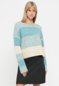 Mladá žena v měkkém pleteném pruhovaném svetr v modré, bílé a šedé barvě, oblečená v černé sukni, stojící před jednoduchým bílým pozadím.