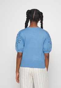 Maglione blu lavorato a maglia con maniche a sbuffo, caratterizzato da motivi tagliati sulle maniche. Indossato con pantaloni a righe leggeri. Vestibilità standard.