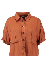 Lyhythihainen, ruosteenvärinen paita, joka on valmistettu sileästä kankaasta. Tässä paidassa on kaulus, nappikiinnitys edessä ja kaksi rintataskua läpillä.