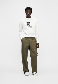 Λευκό πουκάμισο με μακριά μανίκια και μαύρο γραφικό σχέδιο μπροστά, συνδυασμένο με χακί παντελόνι cargo και λευκά αθλητικά παπούτσια.