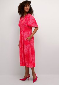 Červené šaty s paisley potiskem, krátkým rukávem, výstřihem do V a bočními kapsami. V kombinaci s růžovými lodičkami na podpatku, zobrazené na neutrálním pozadí.