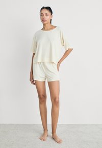 Világos bézs loungewear szett, amely magában foglal egy laza, rövid ujjú, cropped felsőt és egy szoros rövidnadrágot, puha, texturált anyagból készült.