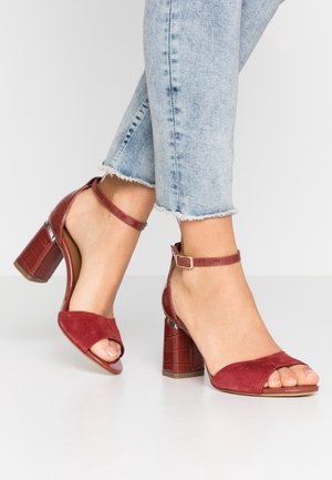 Pieds portant des sandales rouges à talons hauts avec brides à la cheville et talons blocs, associées à un jean bleu clair à ourlet effiloché sur fond de sol blanc.